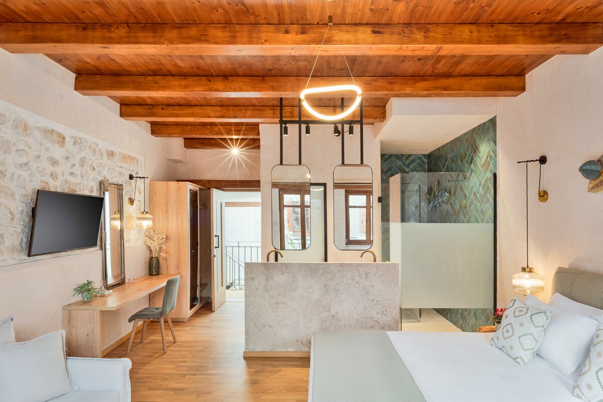 The Superior Suite features wooden ceiling beams and a light wood floor. A spacious bed is accompanied by a minimalist workspace with a chair. The room is divided by a stylish partition leading to the bathroom, showcasing modern design elements and soft lighting.