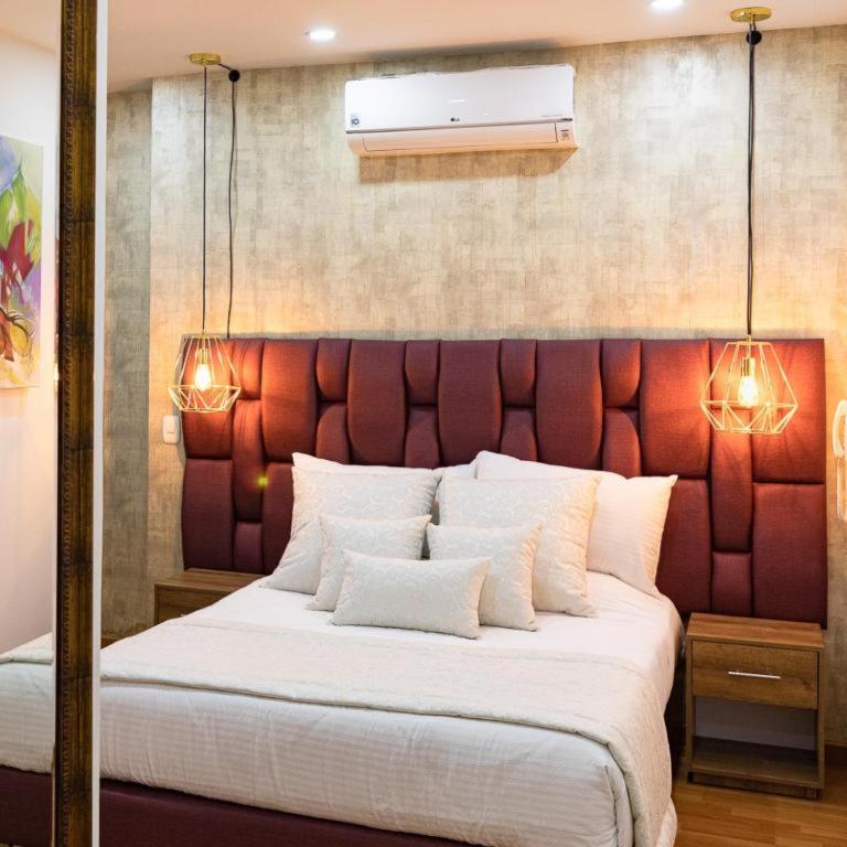 A double room features a plush headboard accentuated by warm wall lighting. Light-colored bedding is complemented by several decorative pillows. Wooden bedside tables provide functional space on either side of the bed. A mounted air conditioning unit is visible above the bed against a textured wall.