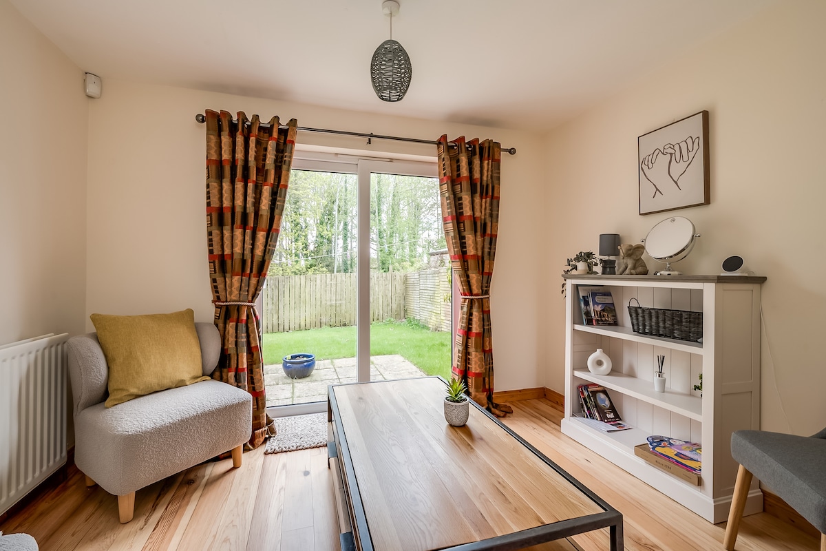 A comfortable living area features soft furnishings, including a light gray sofa and a chair with a vibrant throw pillow. Floor-to-ceiling curtains frame sliding glass doors, which lead to a private outdoor patio area surrounded by greenery. A minimalist bookshelf displays decorative items.