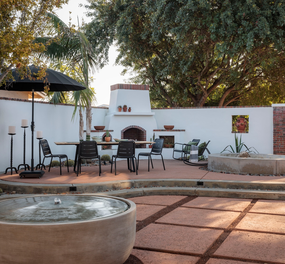 An outdoor patio area features a dining table surrounded by six chairs, a tranquil fountain, and a fireplace with a decorative arch. Verdant trees provide shade, and a large umbrella offers additional coverage. The space is designed for relaxation and gatherings.