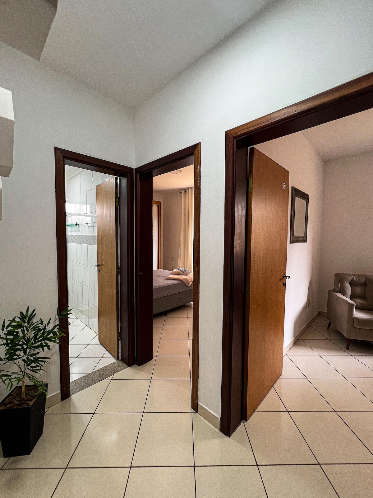 A hallway featuring three doorways leads to different areas of the apartment. A potted plant is positioned beside the entrance. Light-colored walls and tiled flooring create a bright and open atmosphere. One doorway reveals a bedroom with a neatly arranged bed, while another opens to a seating area.