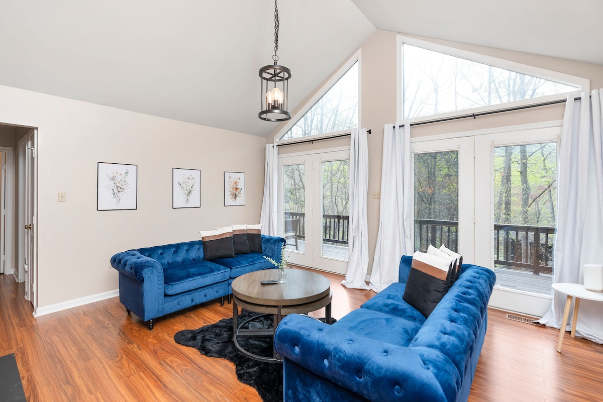A spacious living area features blue velvet sofas arranged around a circular coffee table. Large windows allow natural light to fill the room, with soft white curtains draping beside. Framed botanical prints decorate the walls, complementing the warm wooden flooring.