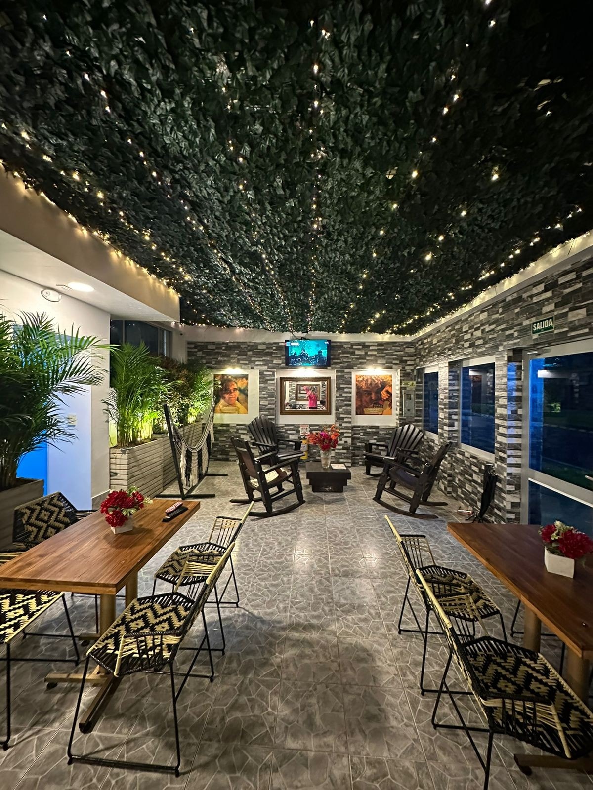 A spacious terrace space is adorned with greenery overhead and warm lighting, creating a relaxed atmosphere. Multiple wooden tables are complemented by patterned chairs. Potted plants add a natural touch, while large windows allow views to the outside, enhancing the inviting feel of the area.