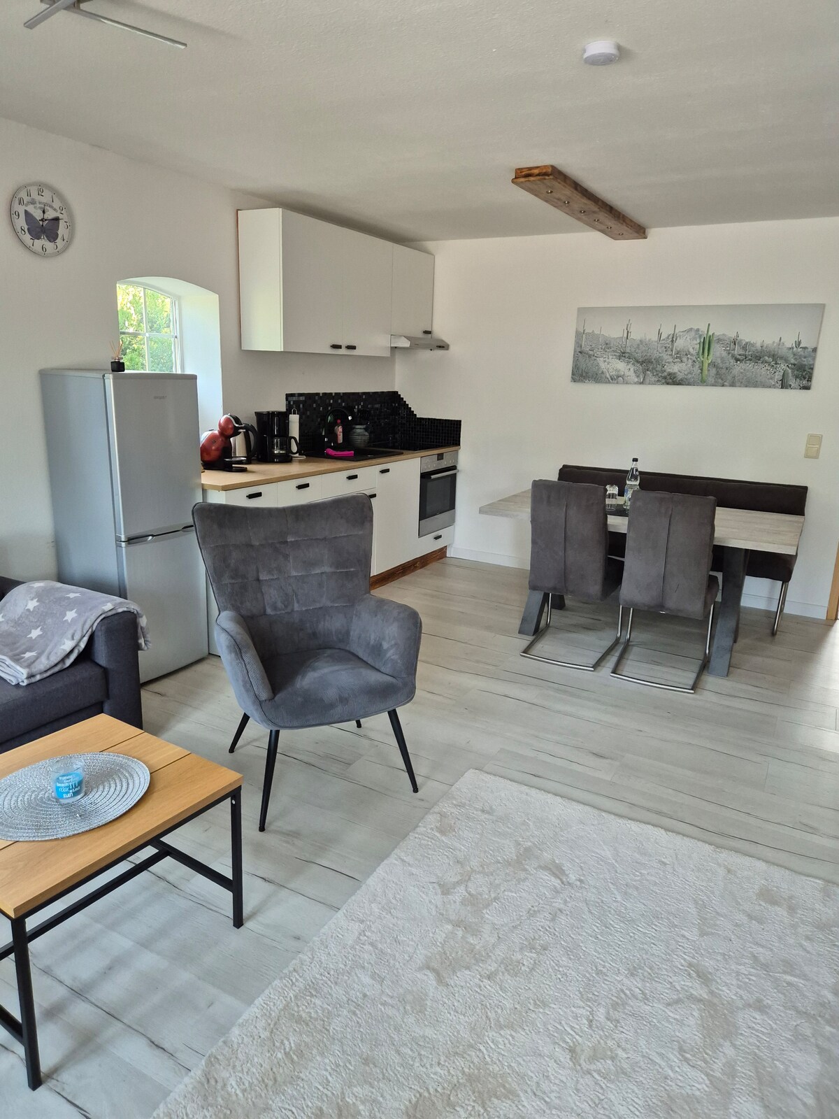 A cozy living area features a modern kitchen equipped with essential appliances and sleek cabinetry. A dining table with four chairs sits adjacent to the kitchen. A comfortable armchair and a small wooden coffee table are positioned in the inviting space, complemented by a light-colored area rug.