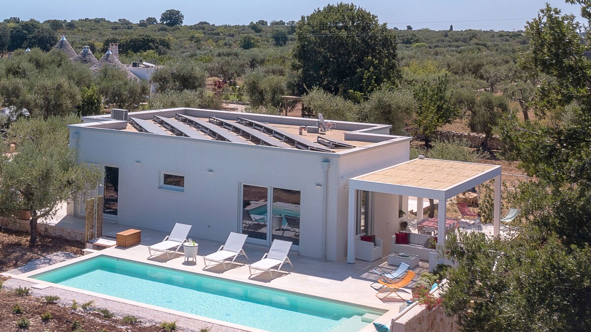 A modern villa is surrounded by lush olive trees and Mediterranean scrub. The swimming pool is accessible from the patio, which is furnished with sun loungers and lounge chairs, creating a serene outdoor space for relaxation.