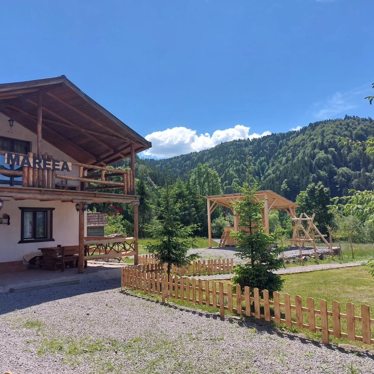 The exterior of the Mareea cabin is displayed under a clear blue sky, showcasing its wooden structure and surrounding greenery. A play area with swings is visible in the yard, along with well-maintained landscaping and a wooden fence bordering the property.