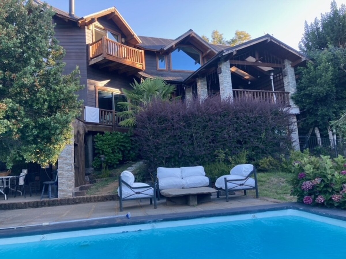 A multi-story wooden house features a stone facade and expansive balconies, surrounded by lush greenery. A refreshing swimming pool is located in front, accompanied by outdoor seating arrangements. The setting reflects a harmonious blend of nature and architecture.