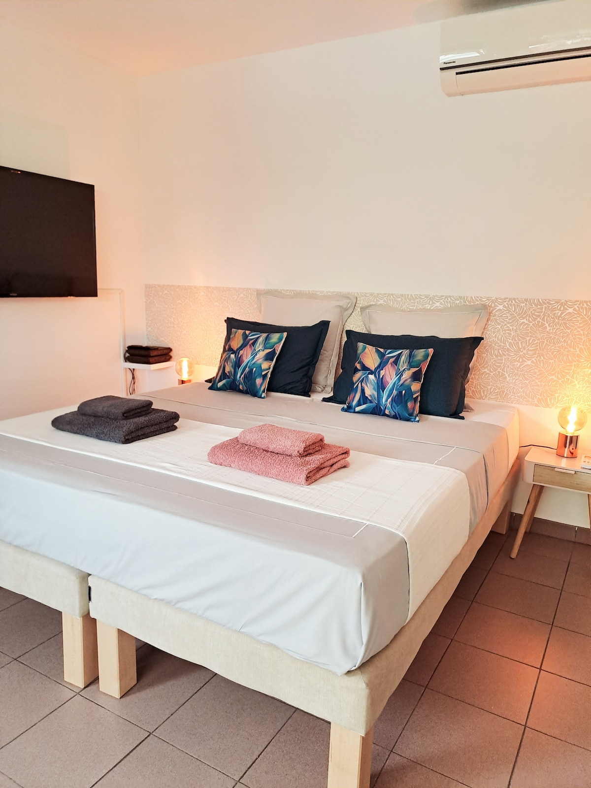 A spacious sleeping area features a king-size bed, adorned with decorative pillows. Soft towels are neatly arranged on the bed. A mounted TV is visible on one wall, with bedside lamps providing gentle lighting. The floor is tiled, enhancing the room's clean aesthetic.
