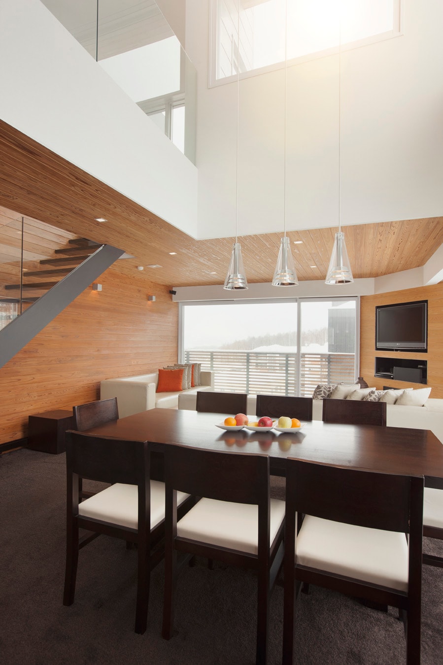 A spacious dining area features a dark wood table surrounded by light-colored chairs, with a bowl of fruit centered on the table. Large windows allow natural light to fill the space, illuminating the contemporary wood and glass design of the room.