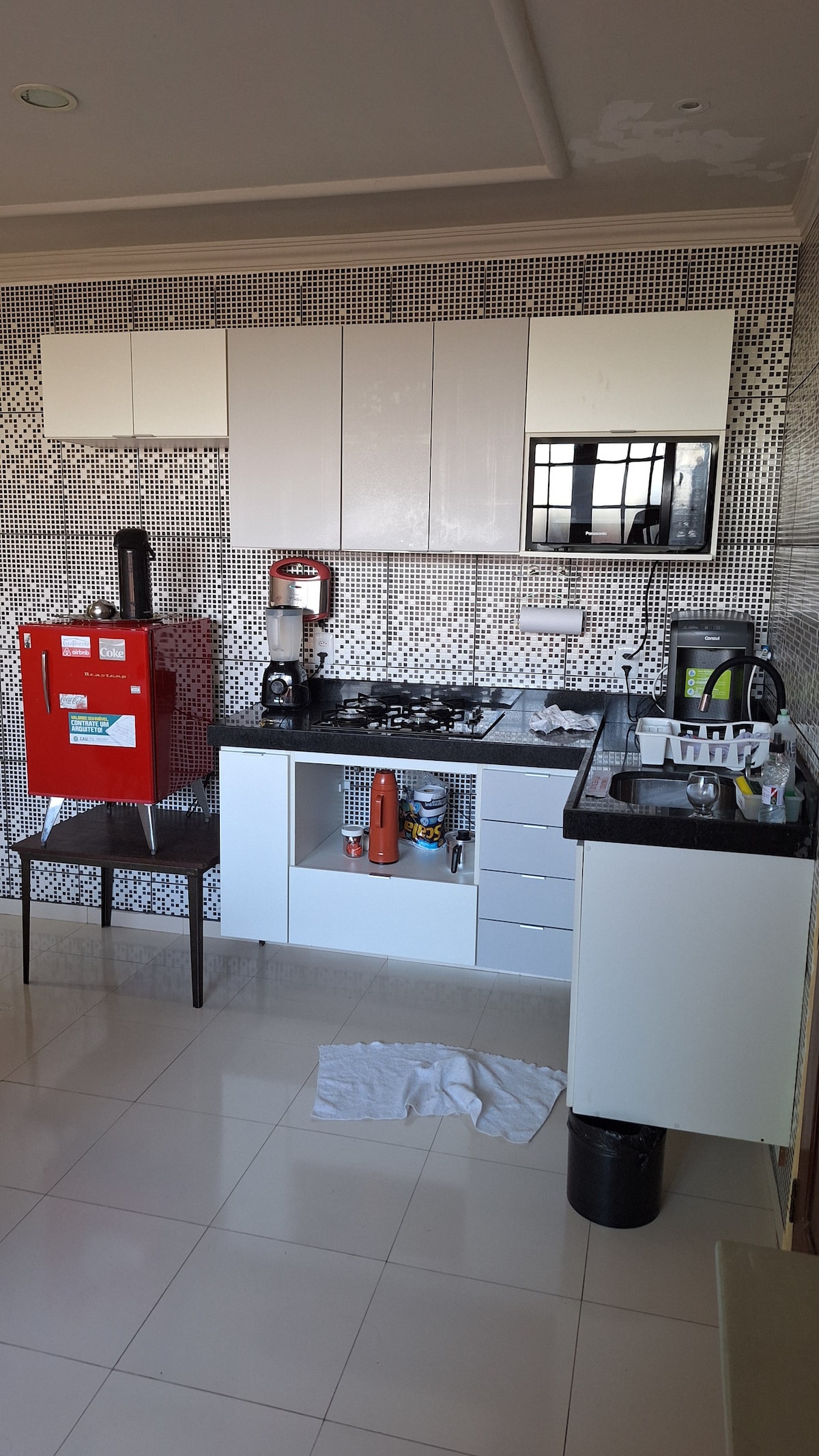 The kitchen features sleek white cabinetry and a mosaic tiled backsplash. Appliances include a red refrigerator, coffee maker, and stovetop. A black countertop provides workspace, while a chair is positioned nearby. Natural light illuminates the area through a window, enhancing the clean, modern aesthetic.
