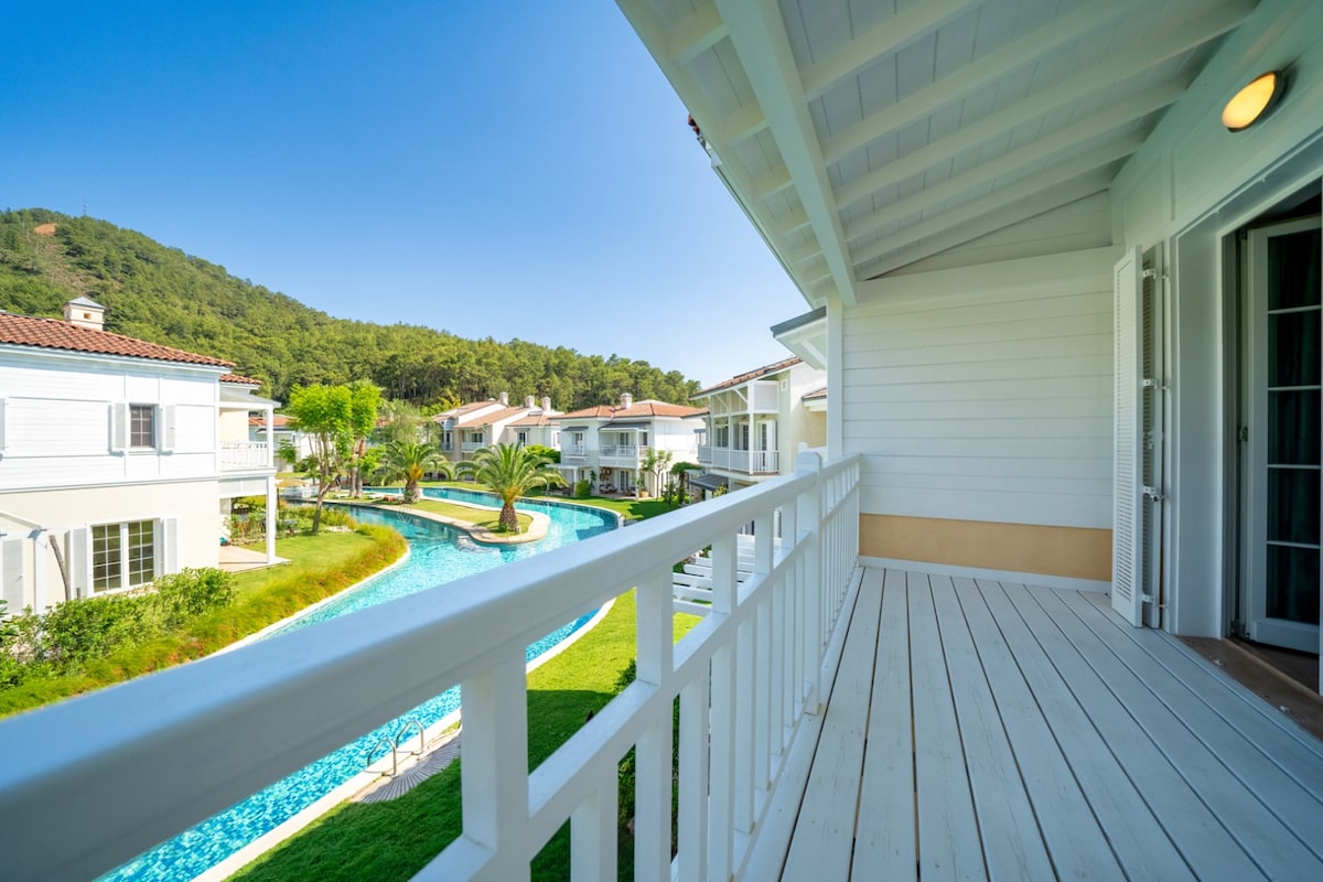 A scenic view of the villa's balcony is captured, featuring a wooden deck that overlooks a winding pool surrounded by lush greenery. The landscape includes palm trees and nearby white buildings, all set against a clear blue sky.