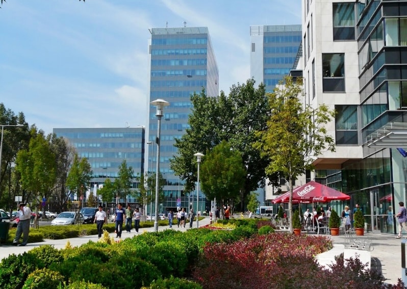 An urban landscape is depicted, featuring modern office buildings with glass facades and green landscaping. People can be seen walking along a pathway, surrounded by shrubs and trees. A shaded outdoor seating area with an umbrella is visible, suggesting a public space for relaxation.
