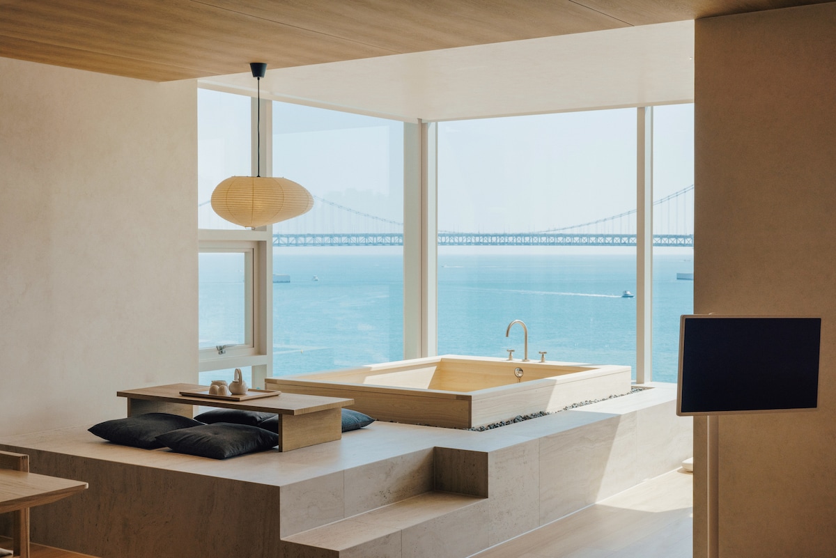 A bright living space features a large window overlooking the ocean, with views of the bridge in the distance. A hinoki bath is positioned by the window, complemented by a dining table and cushions. Natural light enhances the serene atmosphere.