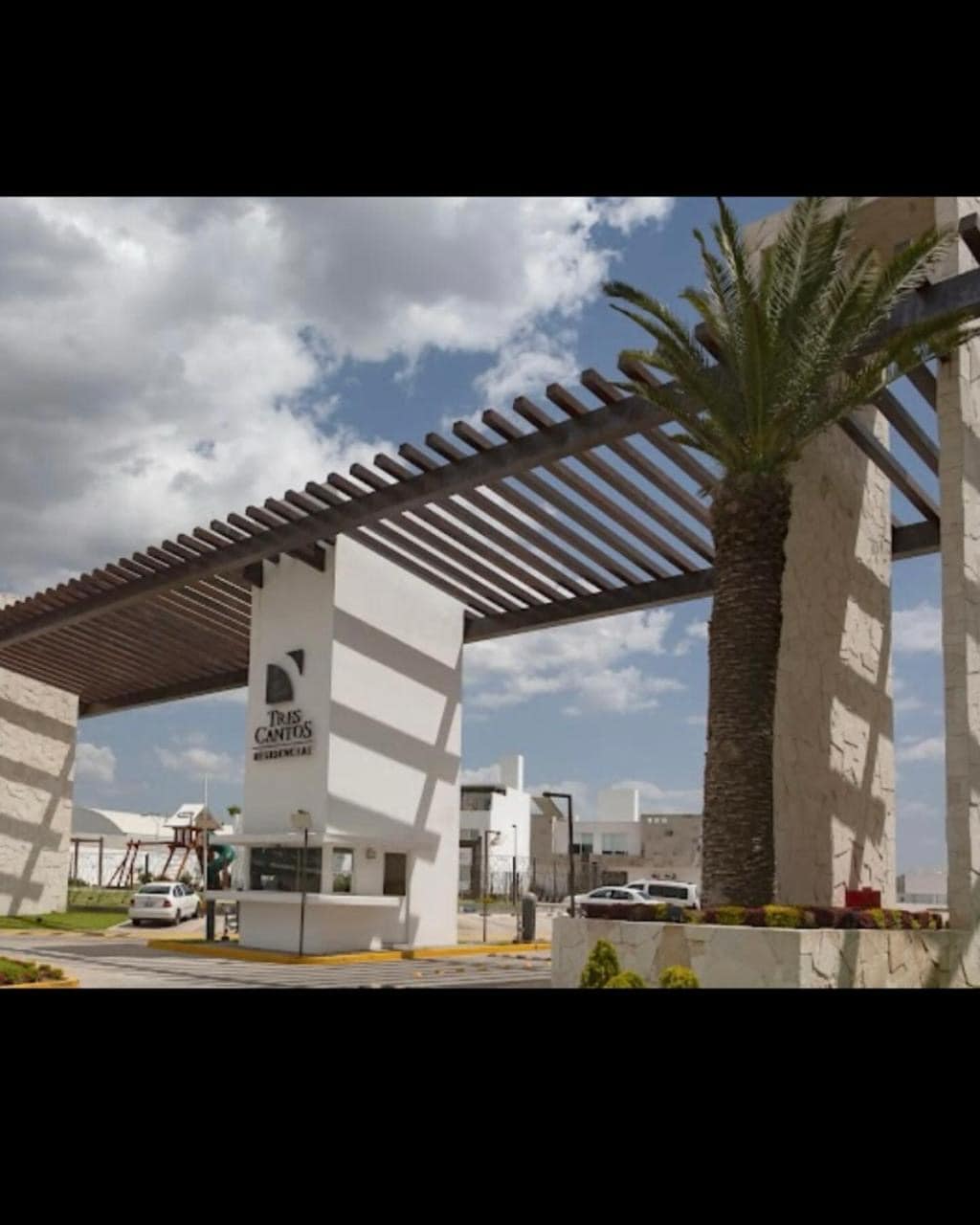 The entrance to the residential area features a modern architectural design with a canopy of wooden beams casting patterned shadows. A palm tree is visible beside the entrance sign, which includes the name of the complex, enhancing the welcoming appearance.