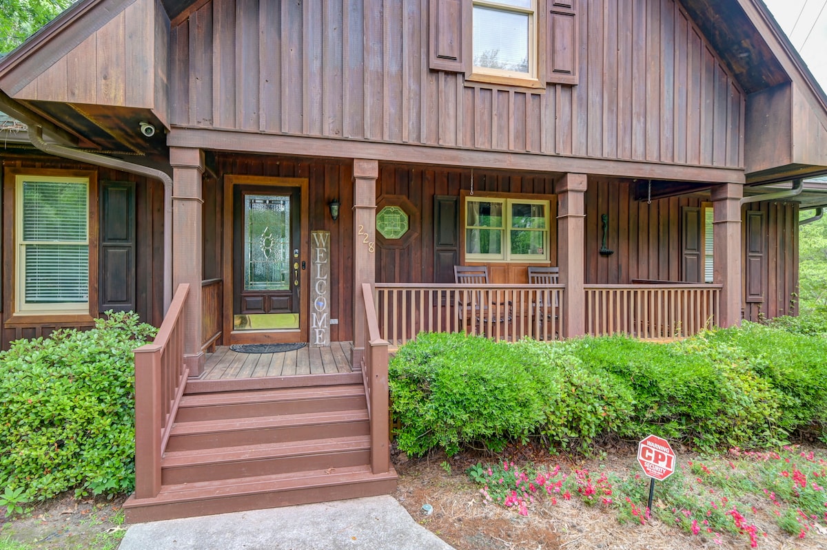 The welcoming entrance features a wooden porch with two rocking chairs. The front door, adorned with a 'Welcome' sign, is flanked by windows that provide natural light. Lush greenery and colorful landscaping enhance the cabin's rustic charm.