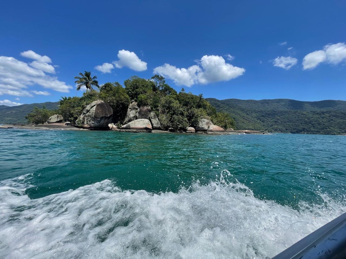 A view of the lush island is captured from a moving boat, showcasing green vegetation and rocky formations along the shoreline. Gentle waves create a frothy trail behind the boat, while a clear blue sky is dotted with white clouds in the background.
