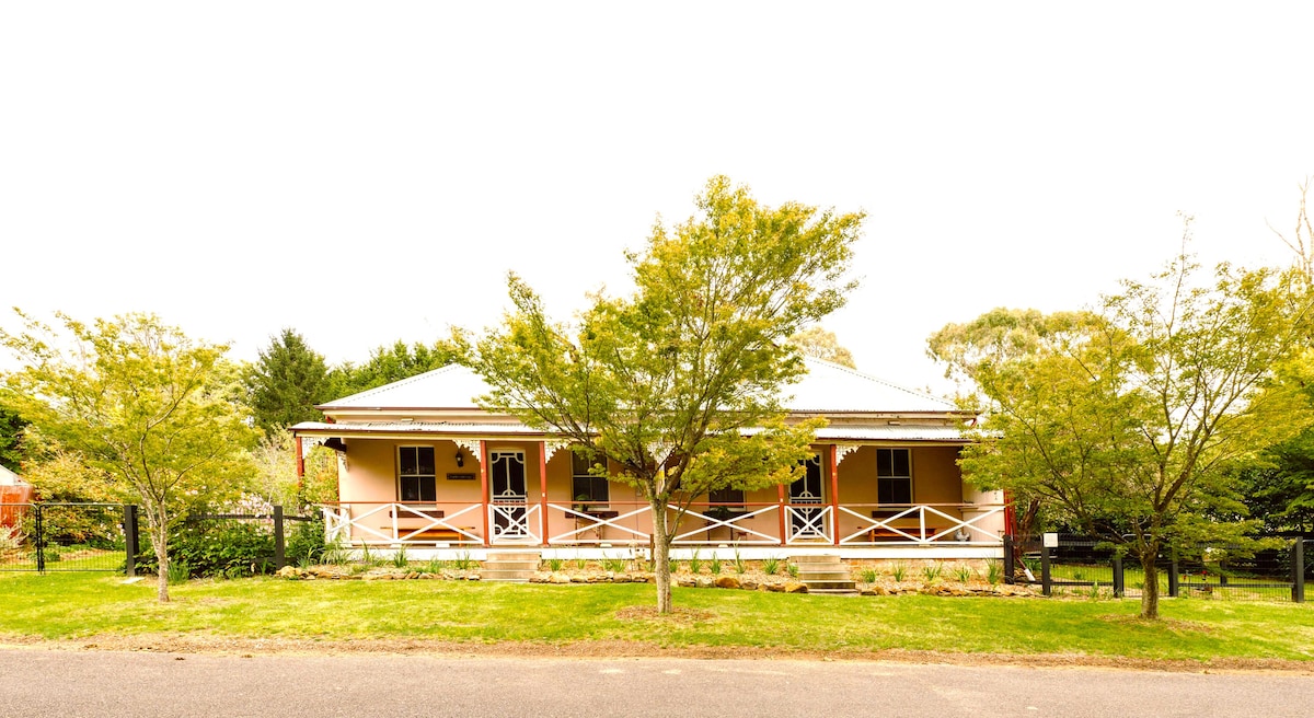 A historic cottage is presented under a clear sky, featuring a welcoming front veranda with white railings. Lush greenery surrounds the property, including neatly trimmed bushes and trees, contributing to a serene ambiance. The façade showcases a traditional style, reflecting its heritage.