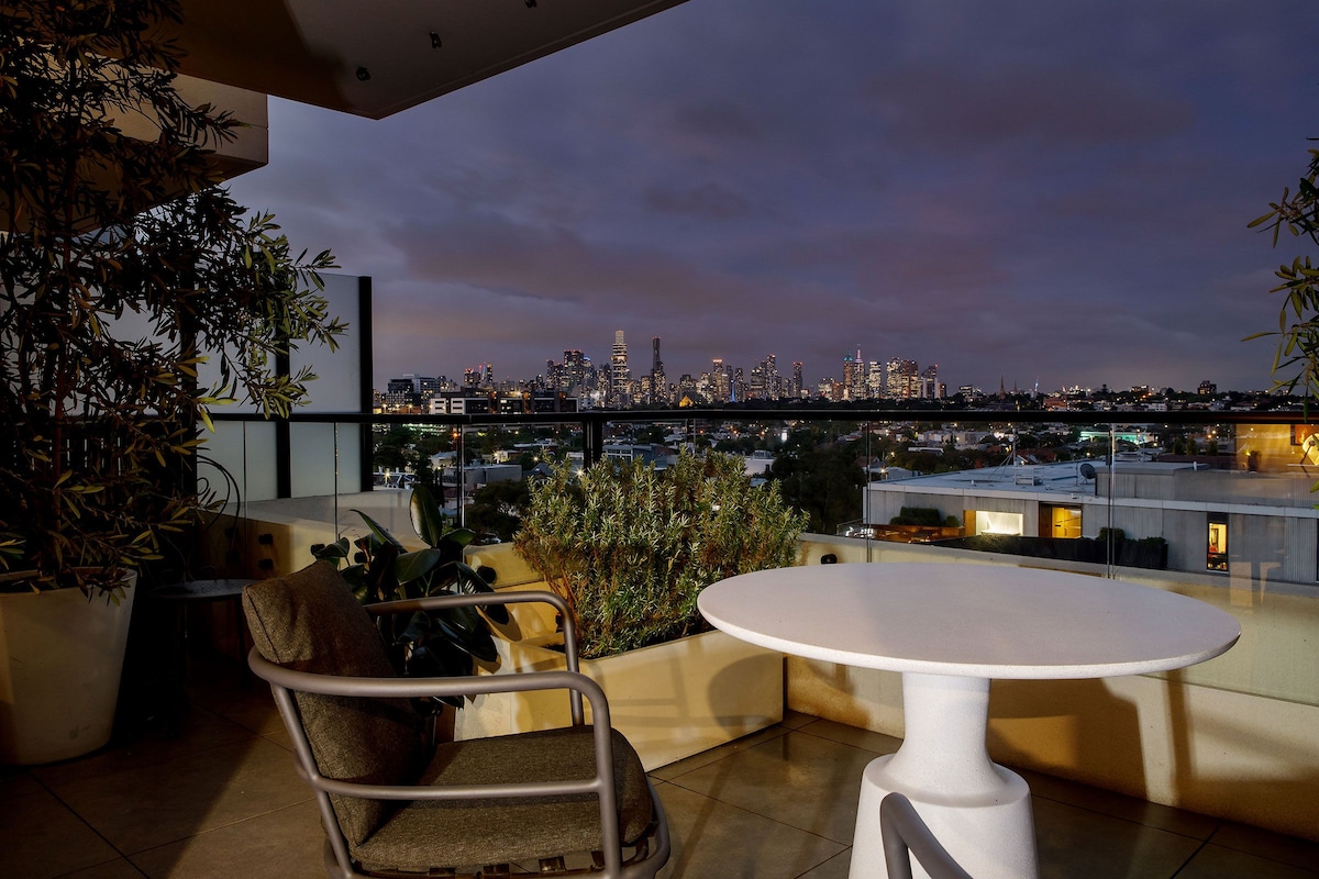 A spacious balcony is presented, featuring a round table and a comfortable chair. Lush greenery surrounds the space, while expansive views of Melbourne's skyline are visible against a twilight sky, showcasing the city's lights beginning to twinkle.