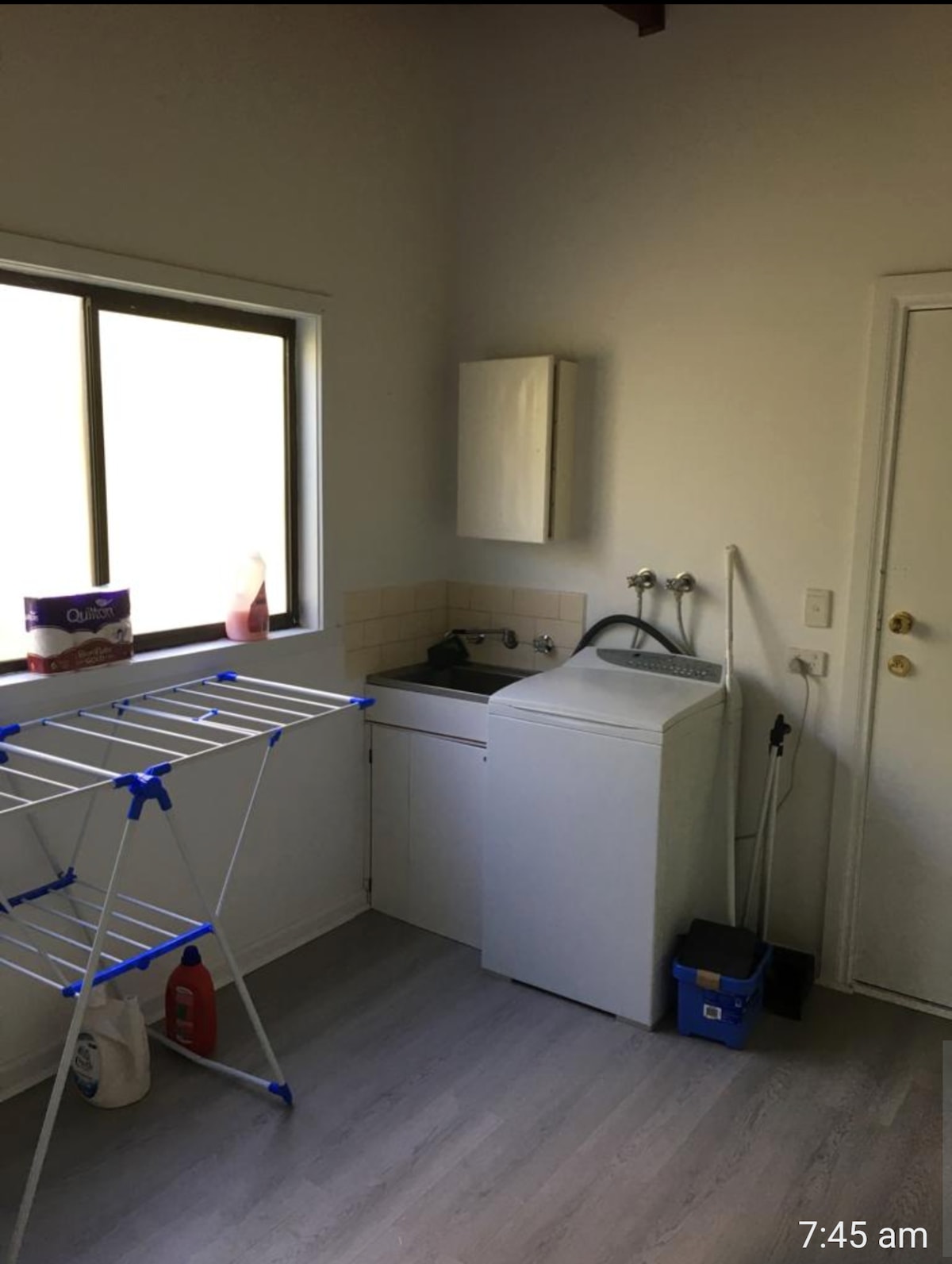 A well-lit laundry area features a washing machine and a sink, with cleaning supplies visible on the countertop. A drying rack stands against the wall near a window, providing natural light to the space. The floor is finished in a light-toned laminate.