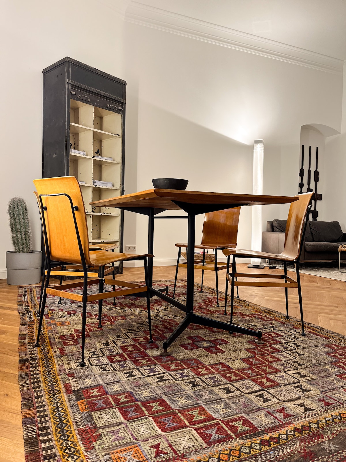 A dining area features a square wooden table surrounded by three chairs. A patterned rug complements the space, while a tall, dark shelving unit stands against the wall, and a modern light source adds gentle illumination. A decorative plant is positioned nearby.