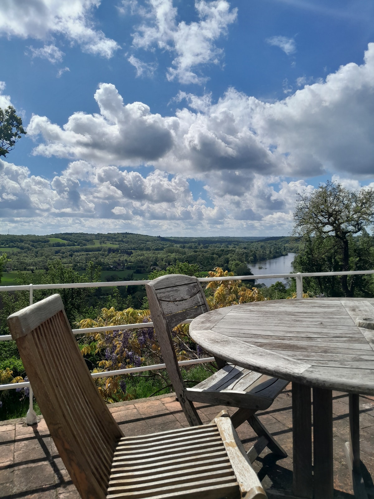 A wooden table and chairs are positioned on a terrace, overlooking a scenic valley with a river. Lush greenery and rolling hills are visible under a partly cloudy sky, creating a peaceful outdoor space for relaxation.