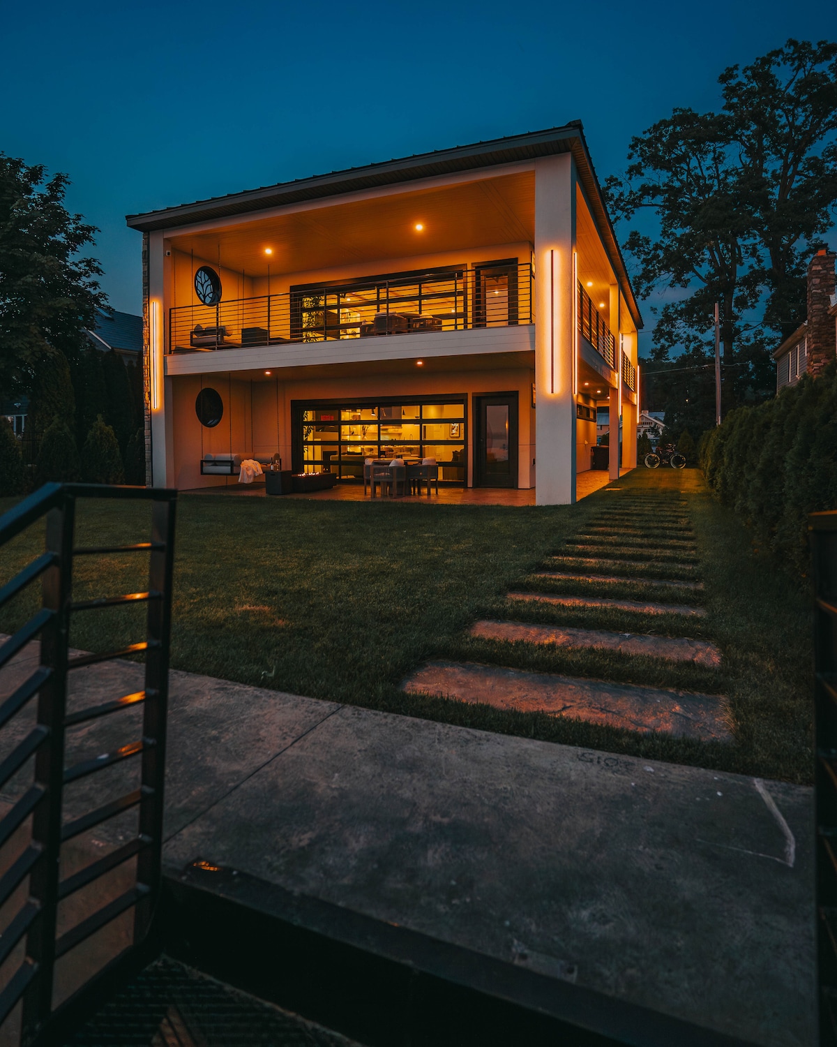 The villa features a modern two-story design, illuminated by warm lighting that highlights its architectural details. A pathway of stone steps leads from the yard toward the entrance, framed by lush greenery and an inviting lawn.