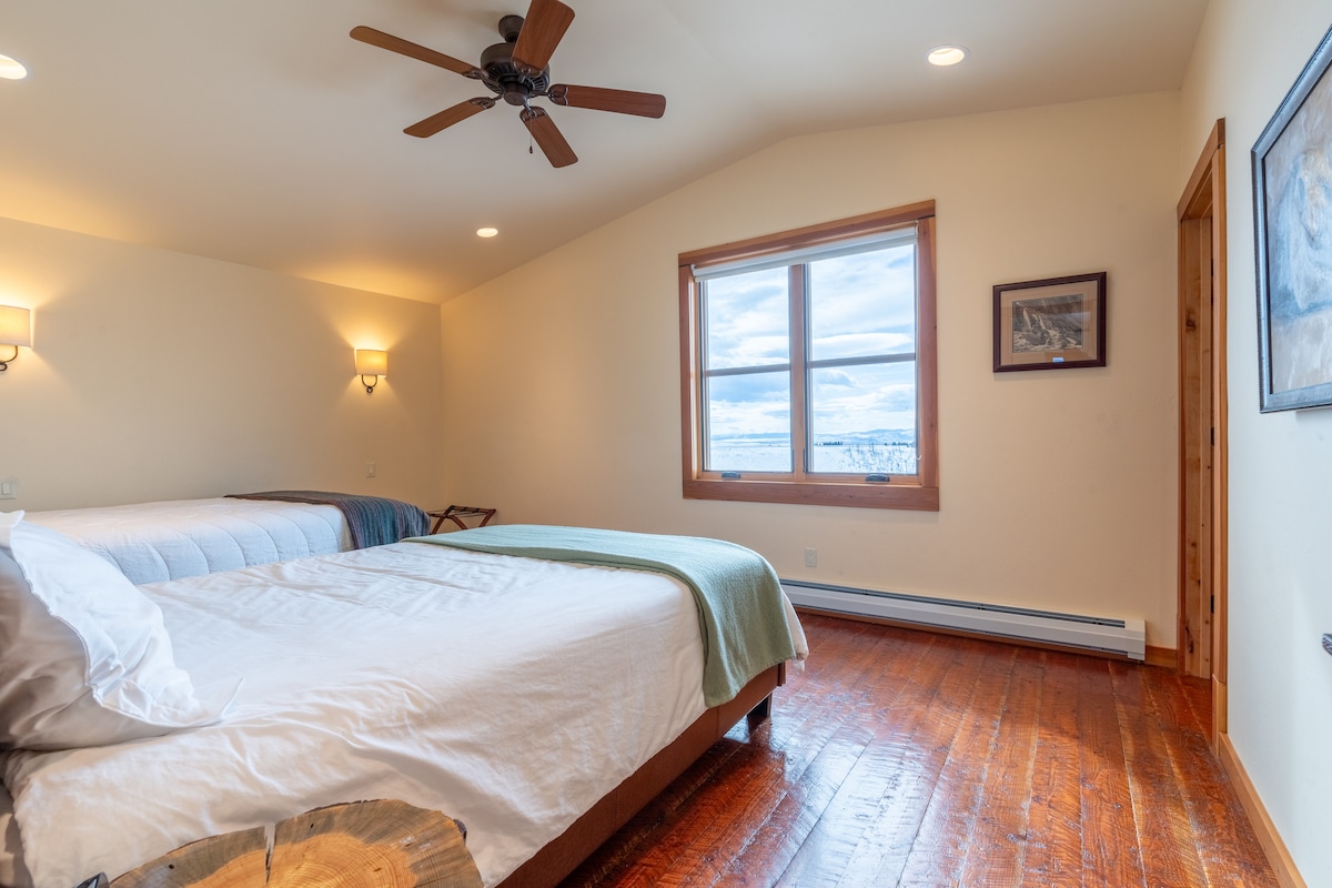 A spacious bedroom features a vaulted ceiling and large windows providing views of the landscape. Two queen beds are dressed in light linens, with a decorative throw on one. Soft lighting is provided by wall sconces on either side of the room.
