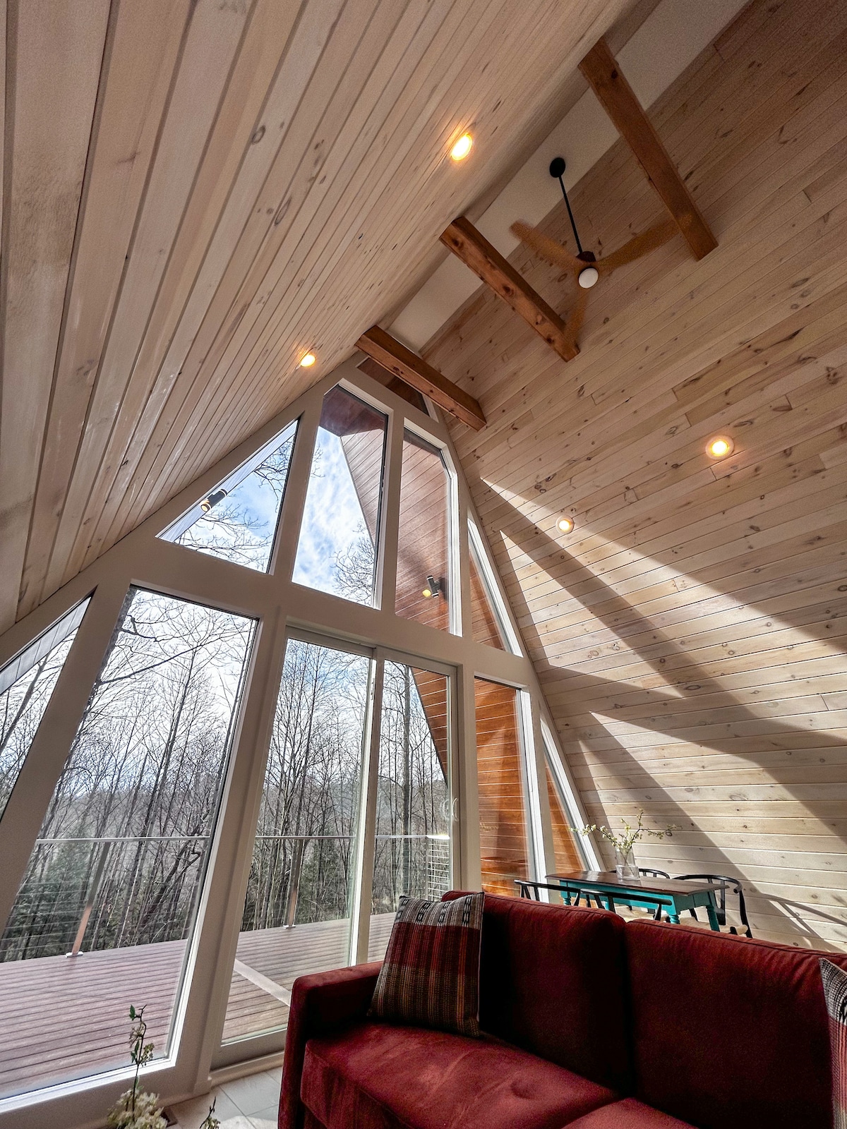 The interior features a modern A-frame design with a high, wooden ceiling and large floor-to-ceiling windows that offer views of the surrounding trees. Soft natural light illuminates the space, while a cozy red sofa adds a touch of comfort.
