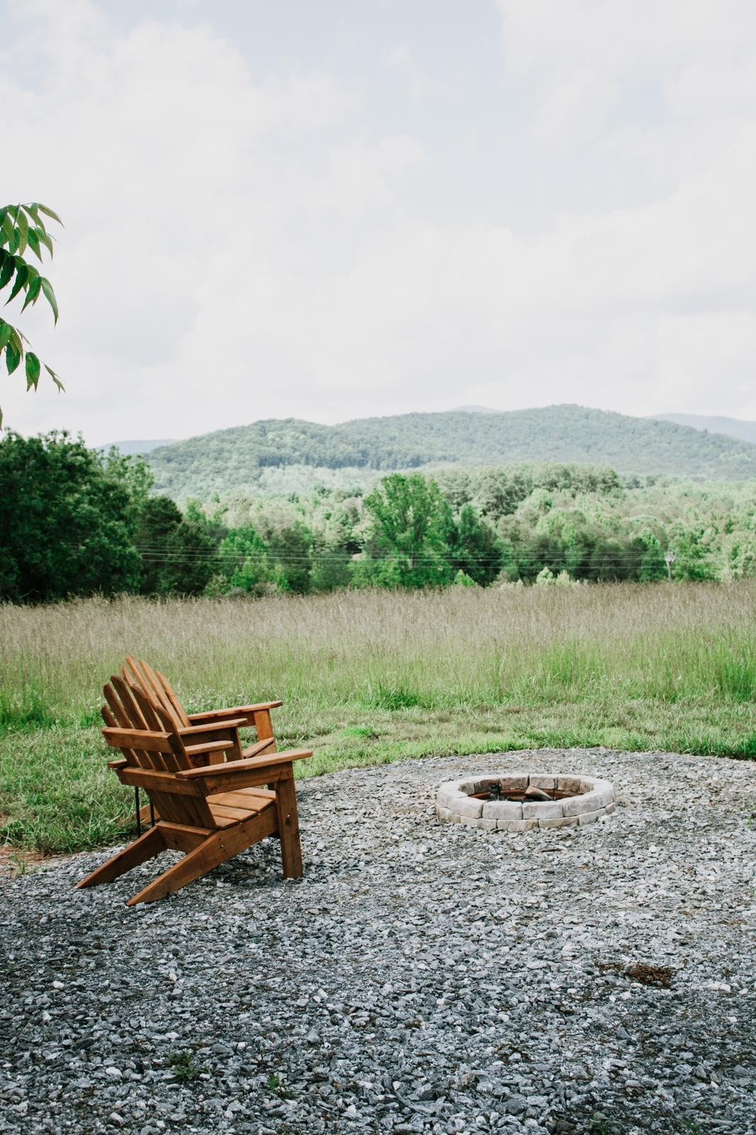 A wooden Adirondack chair is positioned beside a fire pit, surrounded by gravel. In the background, rolling green hills can be seen under a partly cloudy sky, creating a serene outdoor setting.