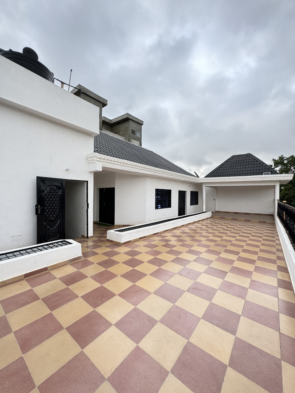 A spacious terrace is captured with a patterned tiled floor in light and dark hues. The terrace features walls painted in white, and various rooftops can be seen in the background under a cloudy sky. Access points are visible, enhancing the space's functionality.