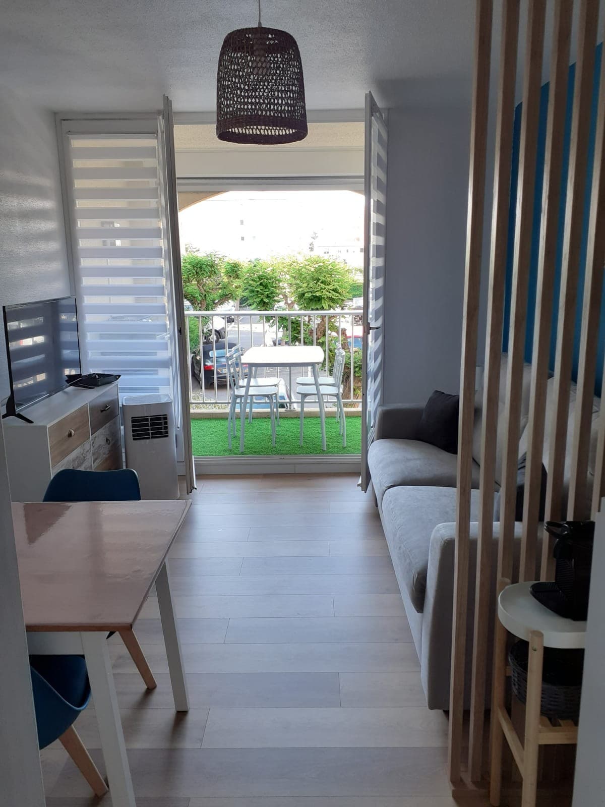 A well-lit living area features a comfortable sofa and a dining space with a table and chairs. Large sliding doors open to a terrace with outdoor seating, surrounded by greenery. A shelving unit adds a touch of separation from the kitchen area.