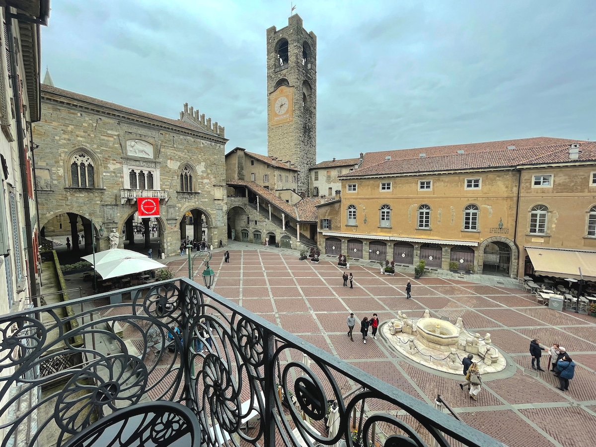 An aerial view of a bustling piazza is showcased, featuring cobblestone paths and historical buildings. A prominent clock tower stands tall, while people stroll around the square. Outdoor seating areas with umbrellas are visible, and a central fountain adds a touch of charm.