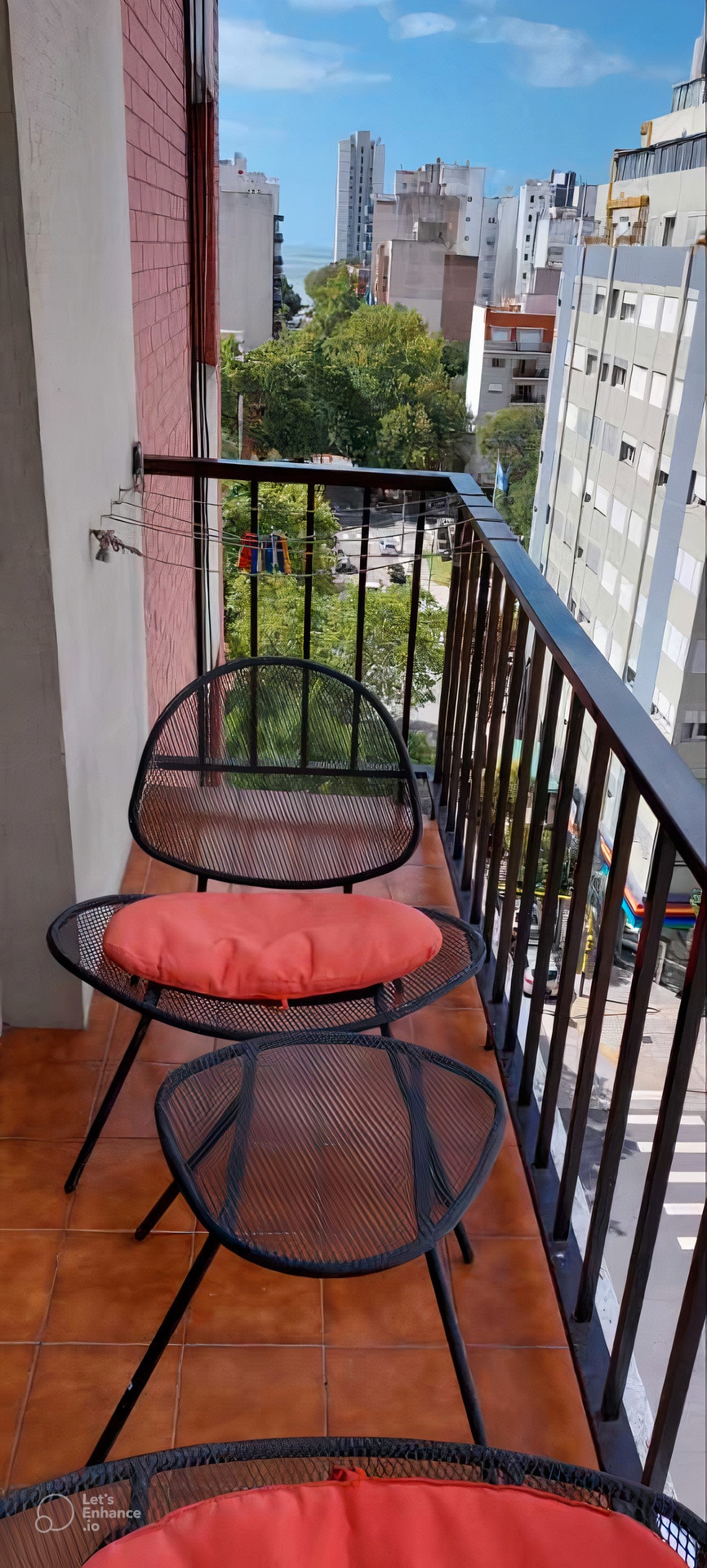 The balcony features four black mesh chairs, each with an orange cushion, arranged against a railing overlooking the city. A view of surrounding buildings and greenery is visible, with bright blue skies enhancing the outdoor space.