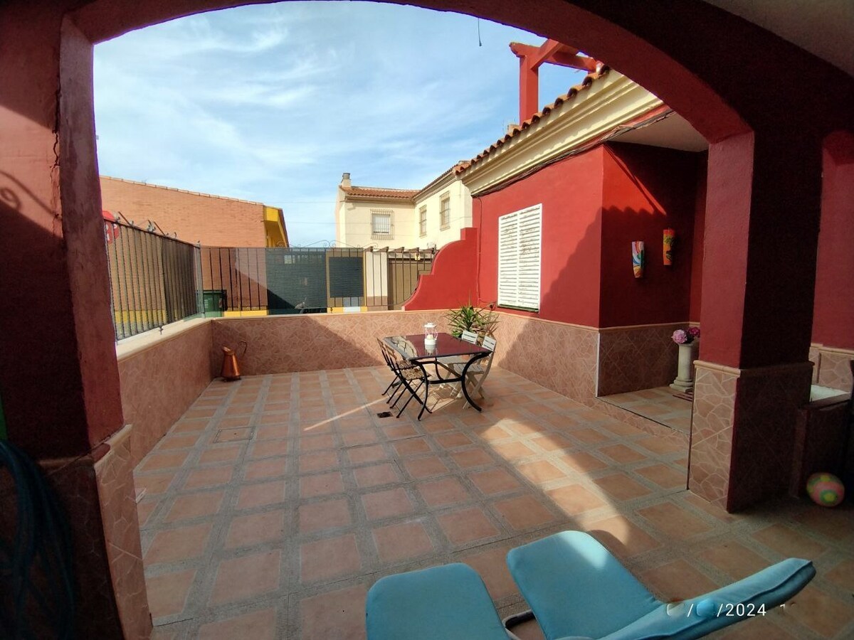 A spacious outdoor area features a tiled floor and an arched entrance. A round table with four chairs is set in the center, surrounded by red walls. Sun loungers are positioned nearby, creating an inviting space for relaxation or dining. Greenery can be seen in the background.