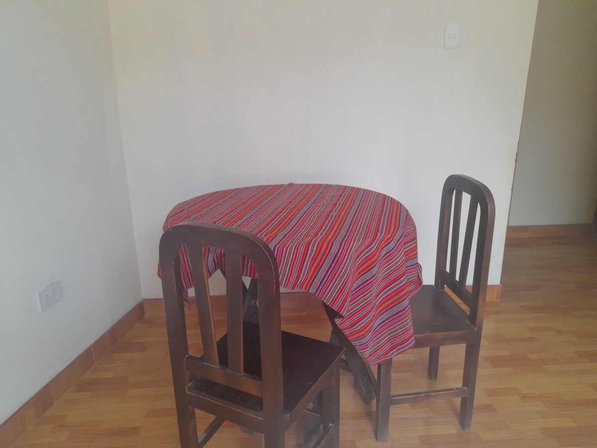 A small dining table with a vibrant striped tablecloth is surrounded by two wooden chairs. The space features hardwood flooring and neutral walls, creating a simple and functional dining area.