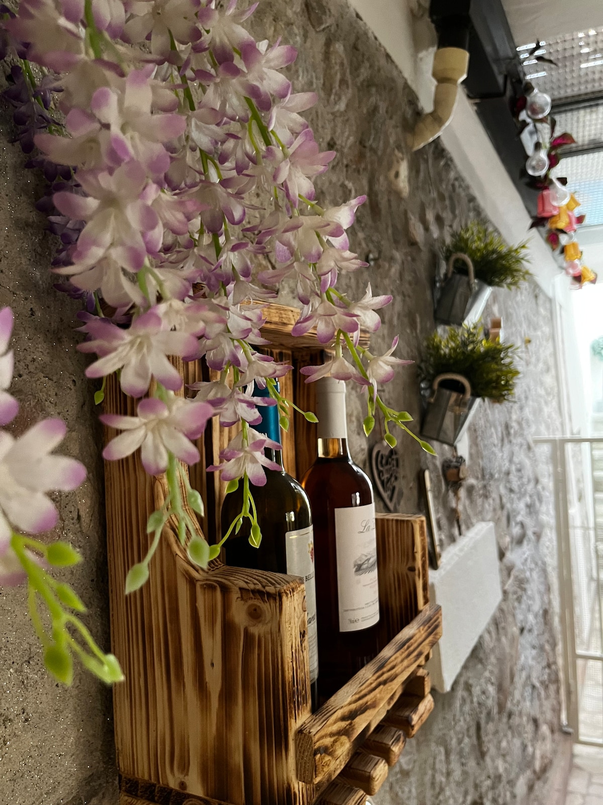 A wooden wine rack is displayed against a stone wall, featuring two bottles of white wine. Delicate, faux purple flowers cascade down from the rack, enhancing the rustic charm of the setting. Small planters with greenery are positioned above the wine rack, adding a touch of vibrancy.