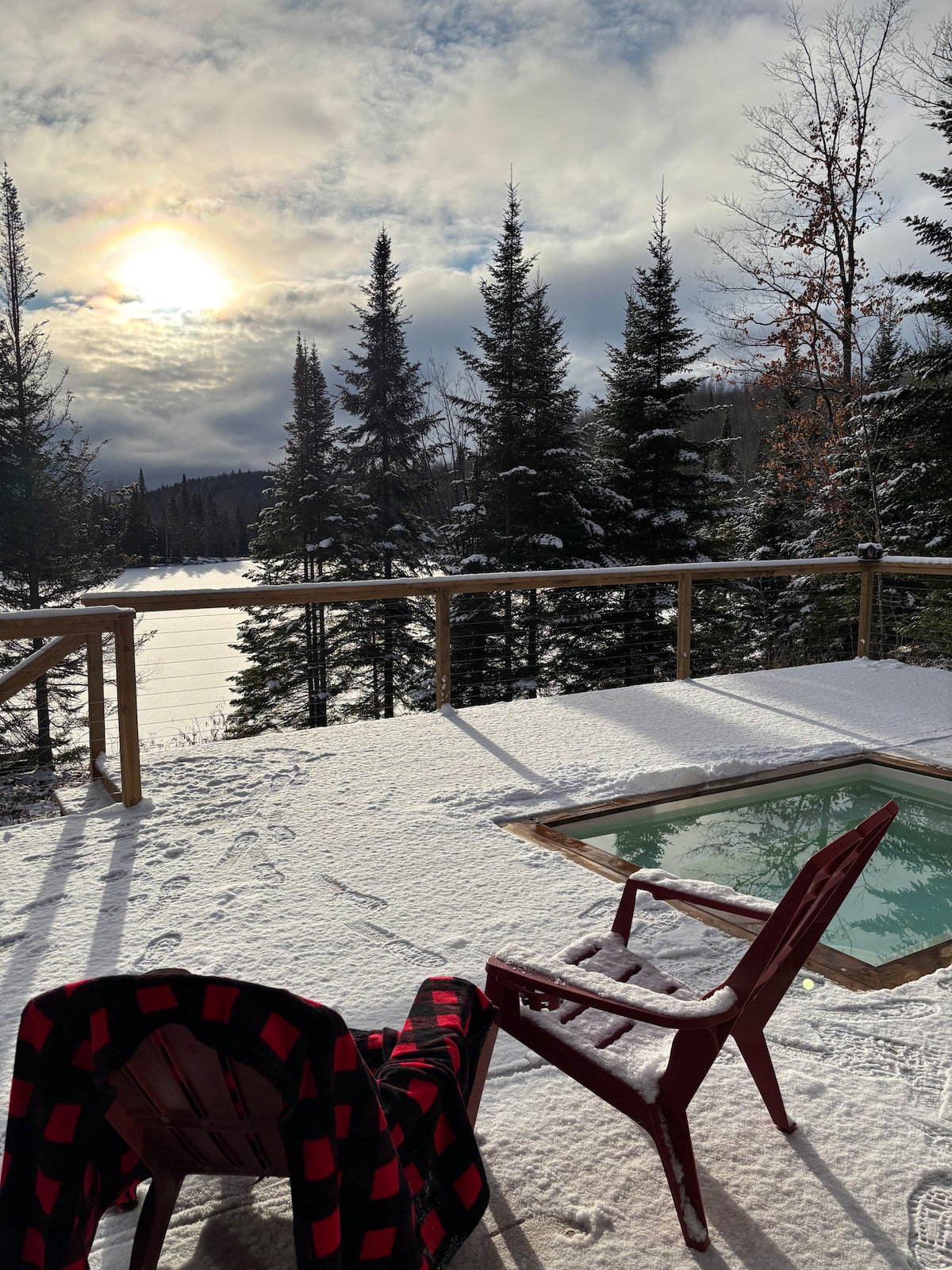 A deck surrounded by evergreen trees is covered in fresh snow, with a view of a tranquil lake in the distance. A cozy red and black plaid blanket drapes over a wooden chair, while sunlight peeks through the clouds, casting a soft glow across the scene.