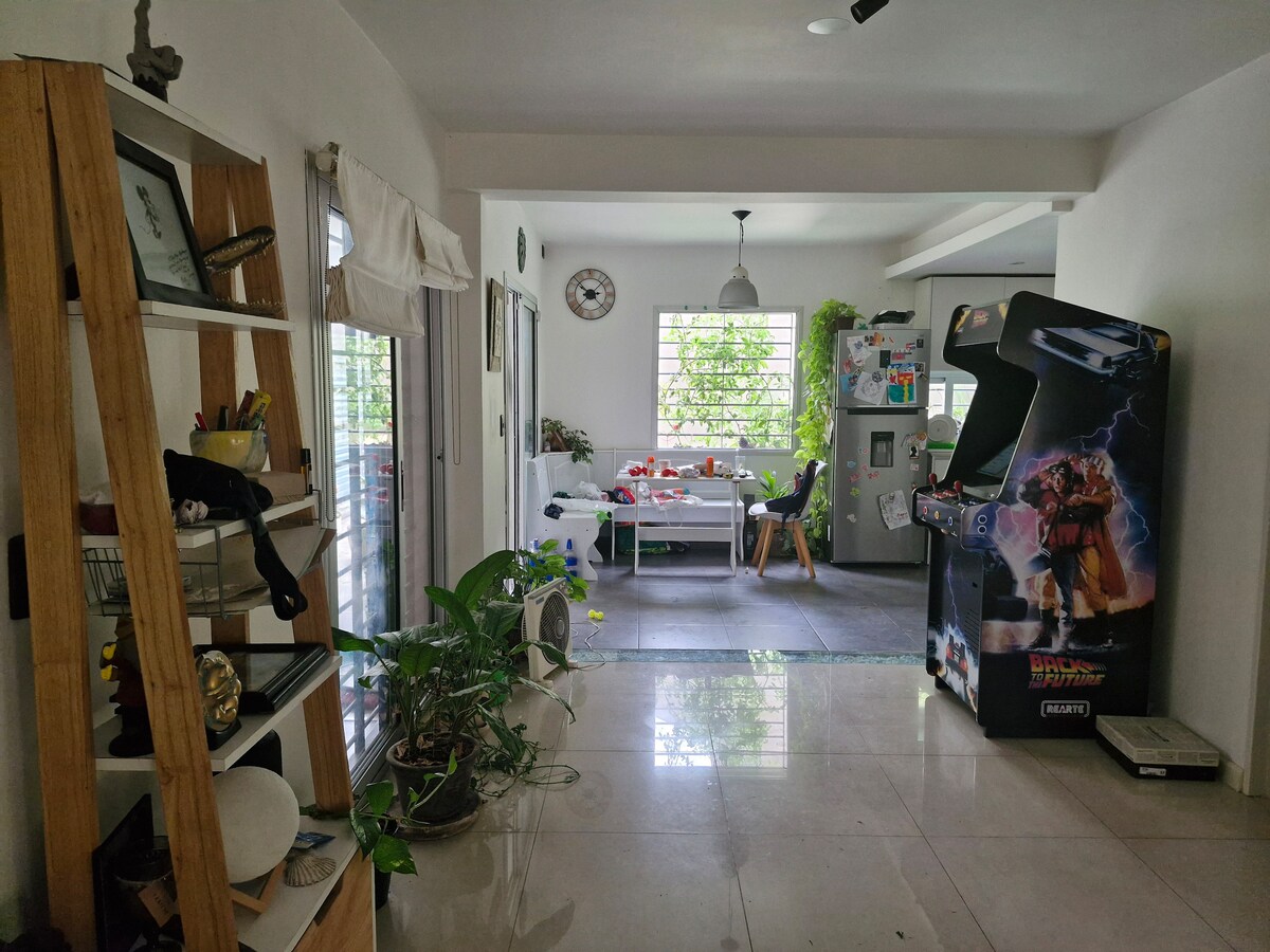 A bright hallway connects two areas of the home. On one side, a wooden shelf is filled with decorative items and plants. A dining area is visible on the opposite side, illuminated by natural light through large windows, with a table set for meals and a vintage arcade machine nearby.