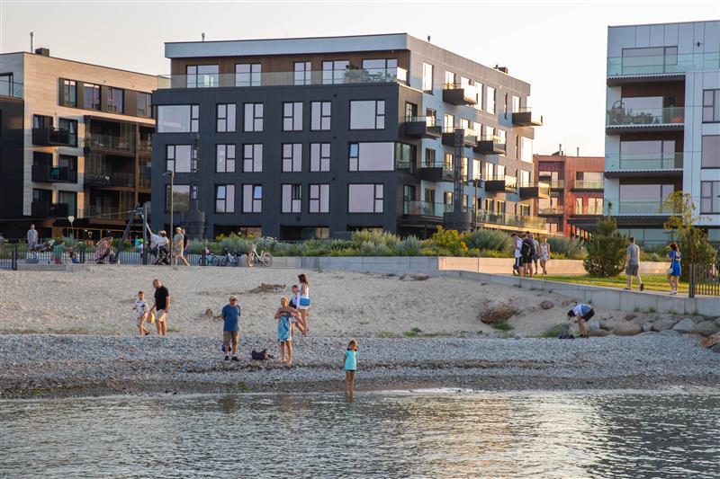 A beach area is shown with families and individuals engaging in leisure activities along the water’s edge. Modern residential buildings are visible in the background, featuring balconies and large windows. The sandy shoreline is complemented by pebbles and greenery, creating a relaxed coastal atmosphere.