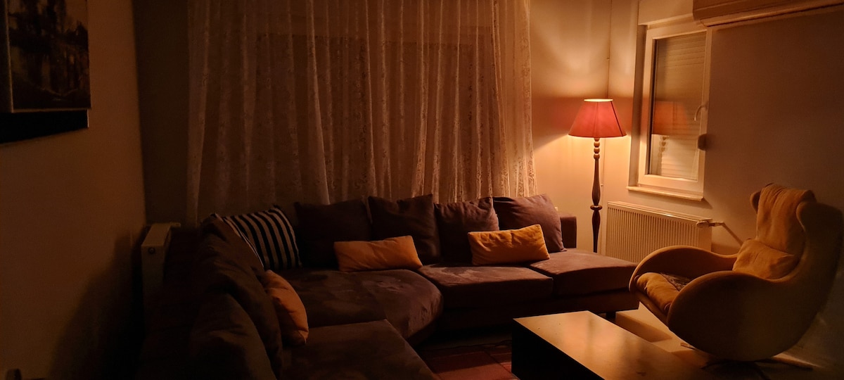 A spacious living area is softly lit, featuring a large sectional sofa adorned with pillows. A tall lamp casts a warm glow, while a plush yellow armchair adds a pop of color. Sheer curtains diffuse the light from the window, creating an inviting ambiance.