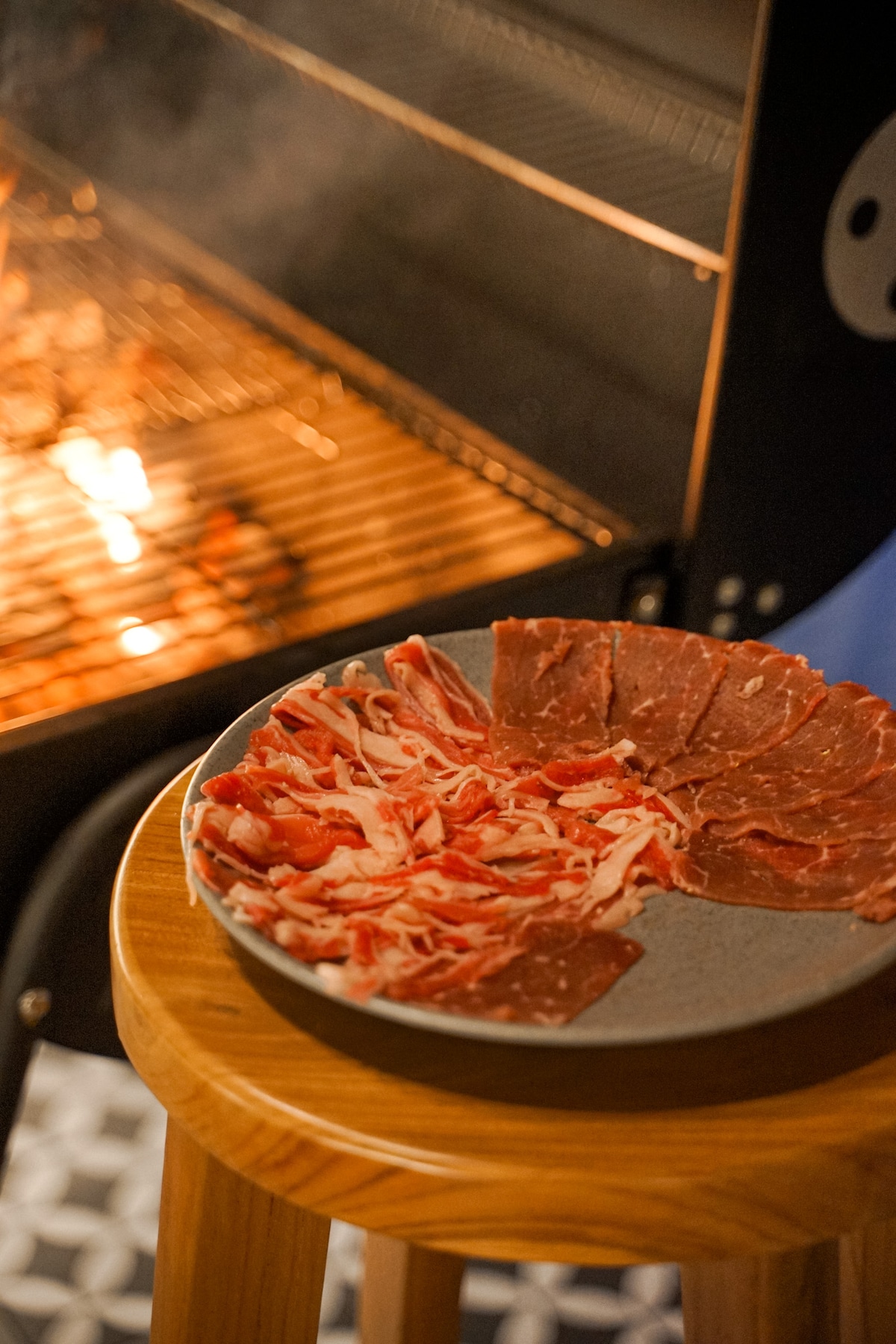 A plate of thinly sliced meat and vegetables is presented on a wooden stool. In the background, a grill emits a soft glow from its heated grates, indicating a cooking process. The warm lighting creates a welcoming setting.