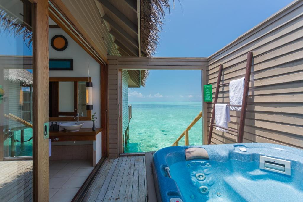 A water bungalow features a private outdoor area with a hot tub overlooking turquoise waters. Large glass doors provide a seamless view of the sea, while natural light illuminates the stylish interior, including a vessel sink and wooden accents.