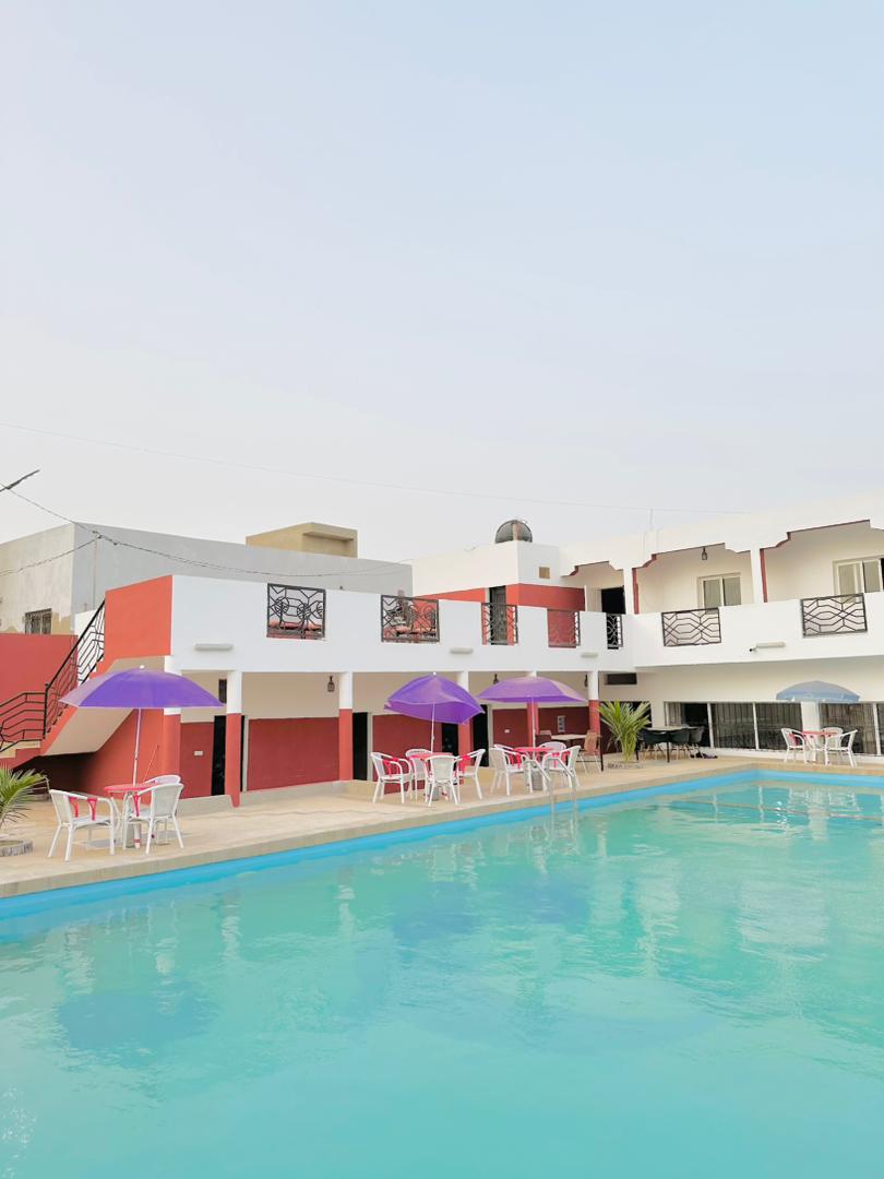 A refreshing pool area is surrounded by a well-maintained patio featuring white tables and chairs. Colorful umbrellas provide shade, while the two-story building showcases a modern design with balconies and large windows. The tranquil water invites relaxation under a clear sky.