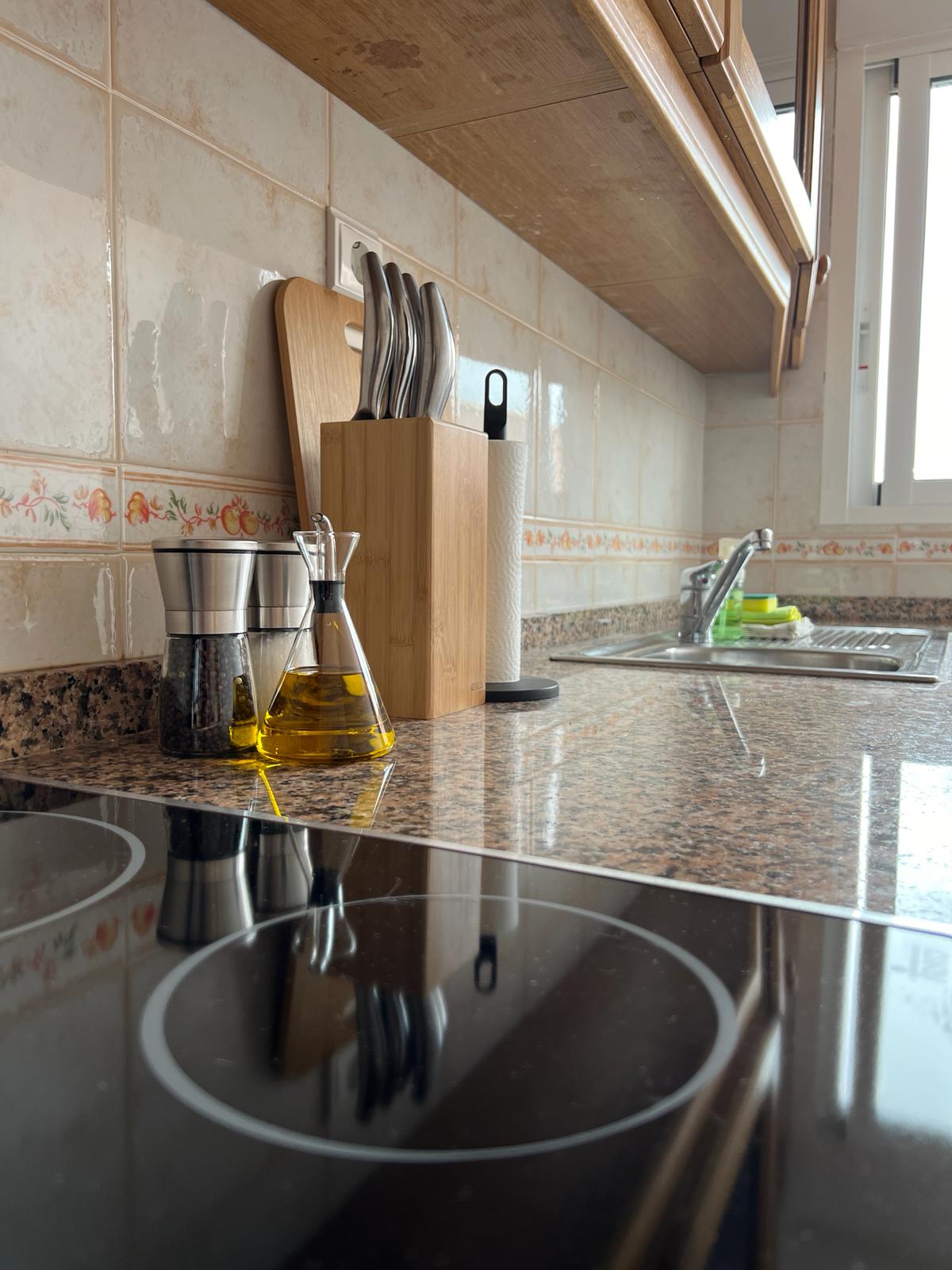 A modern kitchen features a polished countertop with a glass bottle of olive oil and a spice container set nearby. A knife block and utensils are organized on the surface, with a faucet and a glimpse of a window illuminating the space.