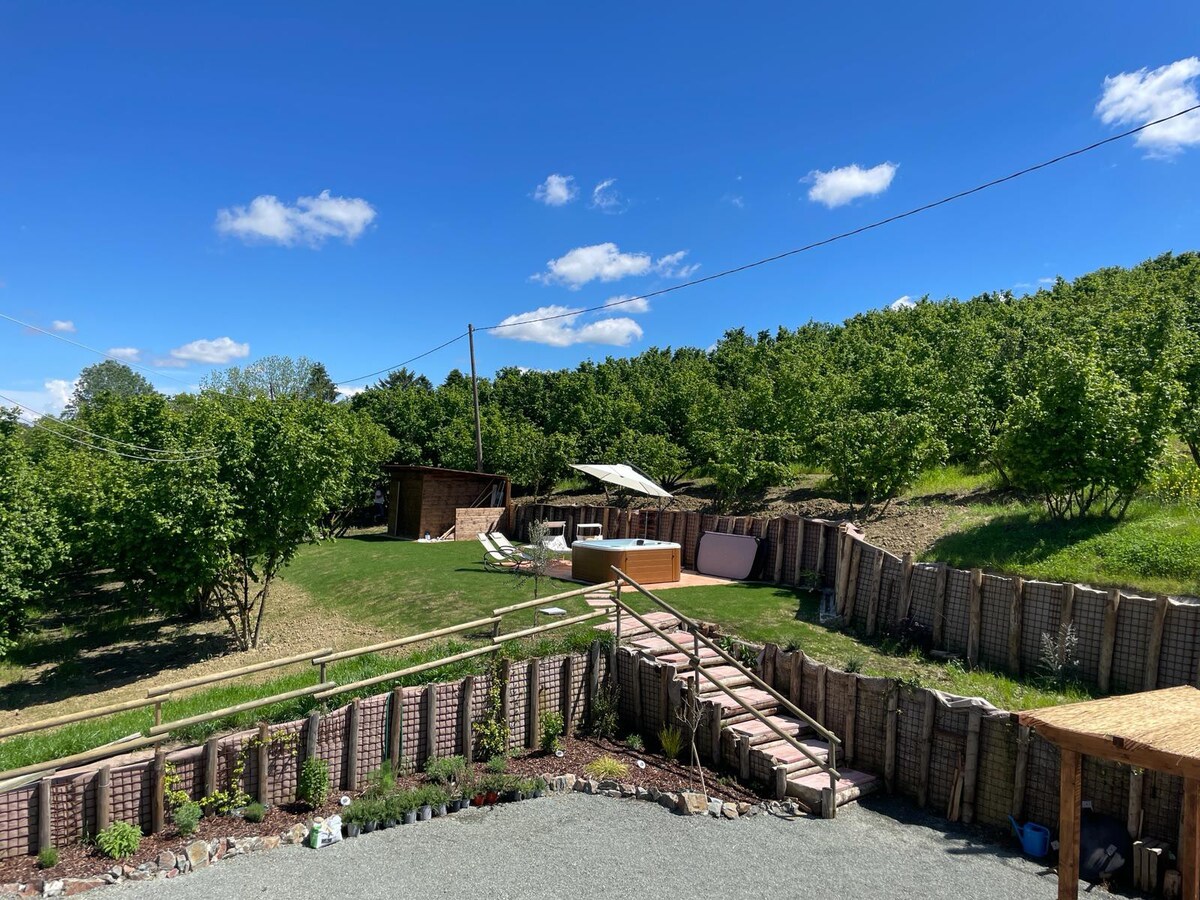 An outdoor space features a well-maintained lawn bordered by trees and greenery. A wooden deck connects the area to a relaxing hot tub and seating. Scenic views of the surrounding nature create a serene setting for guests to unwind.