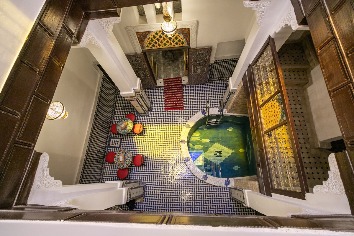 An aerial view captures a spacious courtyard featuring a tranquil pool surrounded by decorative tiles. Red seating is arranged around a central table, while ornate archways and rich wood accents frame the space, highlighting a blend of traditional and contemporary design.