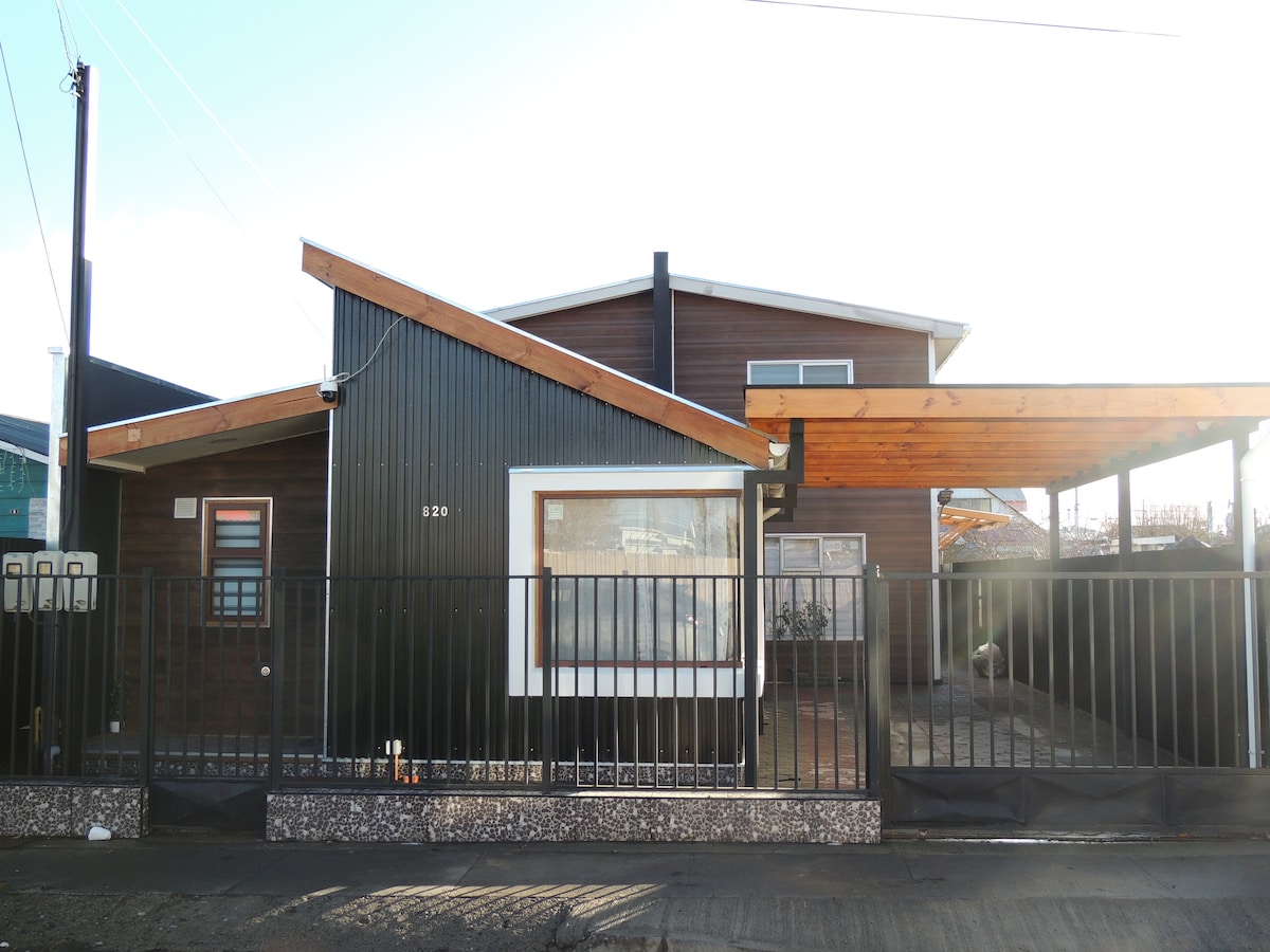 The exterior of the cabin is shown, featuring a modern design with a combination of wooden and metal elements. A gabled roof extends over the entrance, and a dark fence encompasses the property. The surrounding area is well-lit, providing a welcoming appearance.