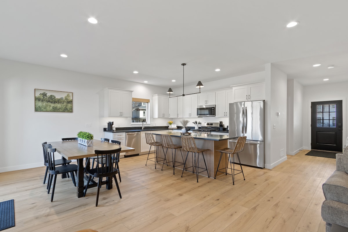 An open concept kitchen and dining area displays modern furnishings. The kitchen features stainless steel appliances, including a refrigerator and gas range. A large dining table with black chairs sits adjacent to a spacious island with seating. Natural light brightens the wooden flooring and walls.