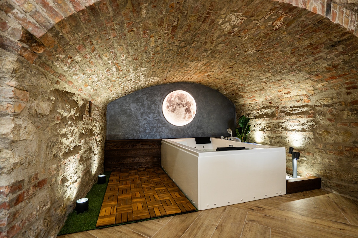 A stylish Jacuzzi is positioned within a natural stone cave, beneath an arched brick ceiling. Soft lighting illuminates the space, highlighting the wooden flooring and a serene circular window that depicts a moon. Greenery adds a touch of nature to the ambiance.