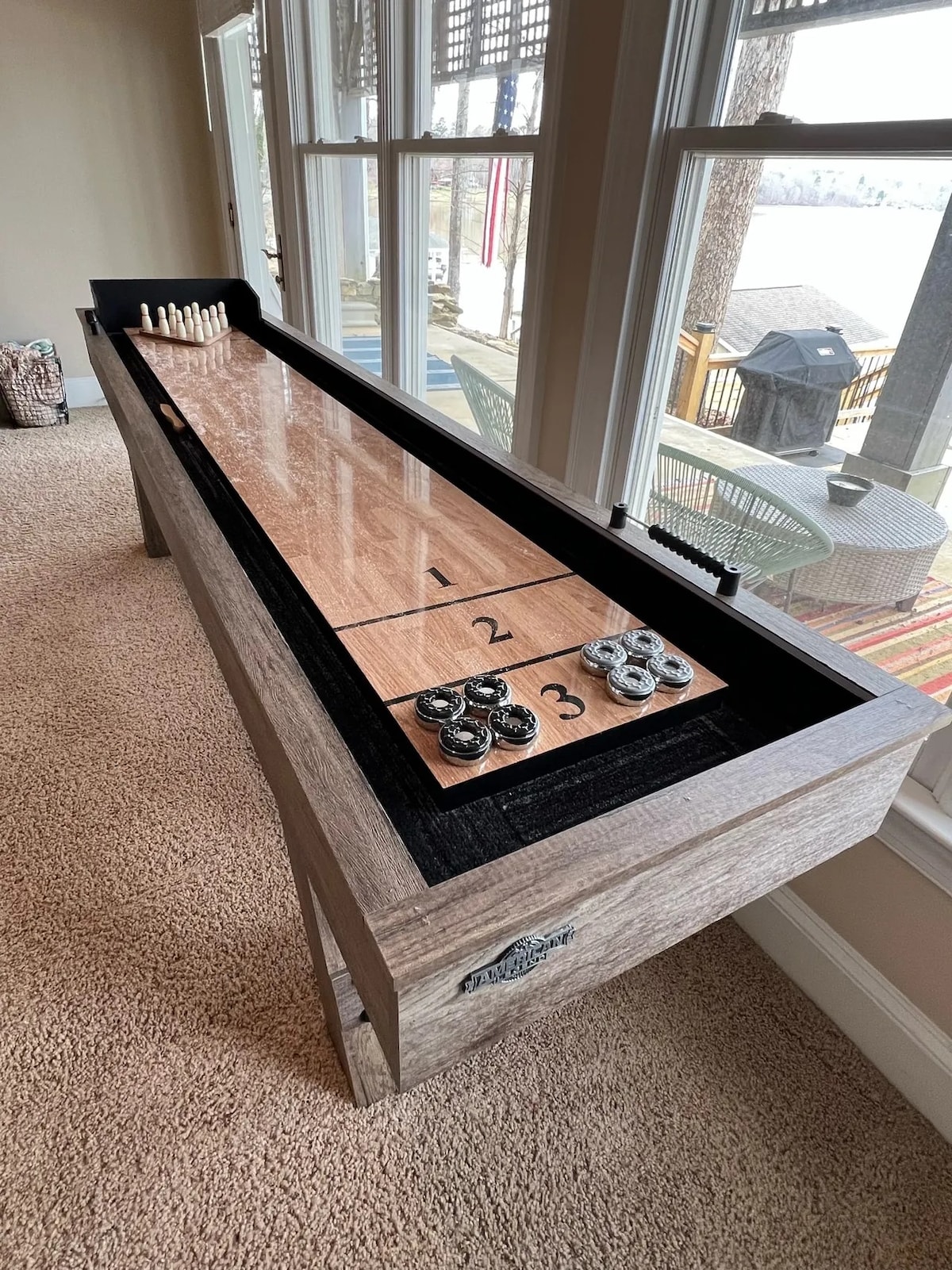 An indoor shuffleboard table is shown, positioned beside large windows that offer views of the lake. The table features a smooth wooden surface with marked scoring sections and an area for placing pucks. Soft carpet covers the floor beneath the table.